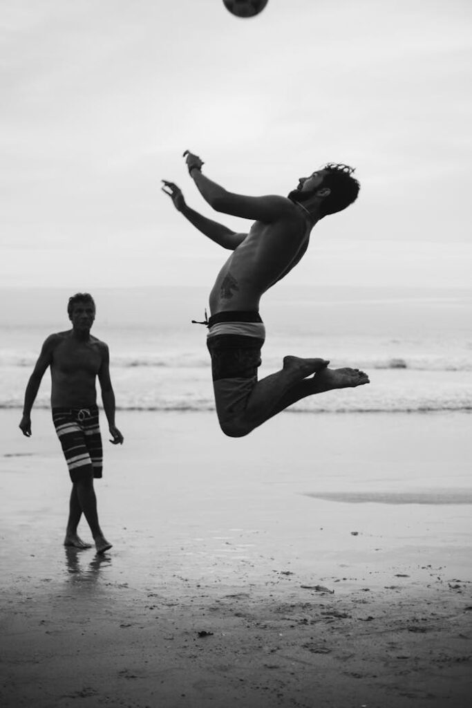 Dynamic shot of a shirtless man leaping to hit a soccer ball on a beach, capturing athleticism and seaside leisure.